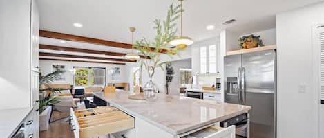 Una cocina moderna con armarios blancos, una isla central con cajones, iluminación colgante y un refrigerador de acero inoxidable.