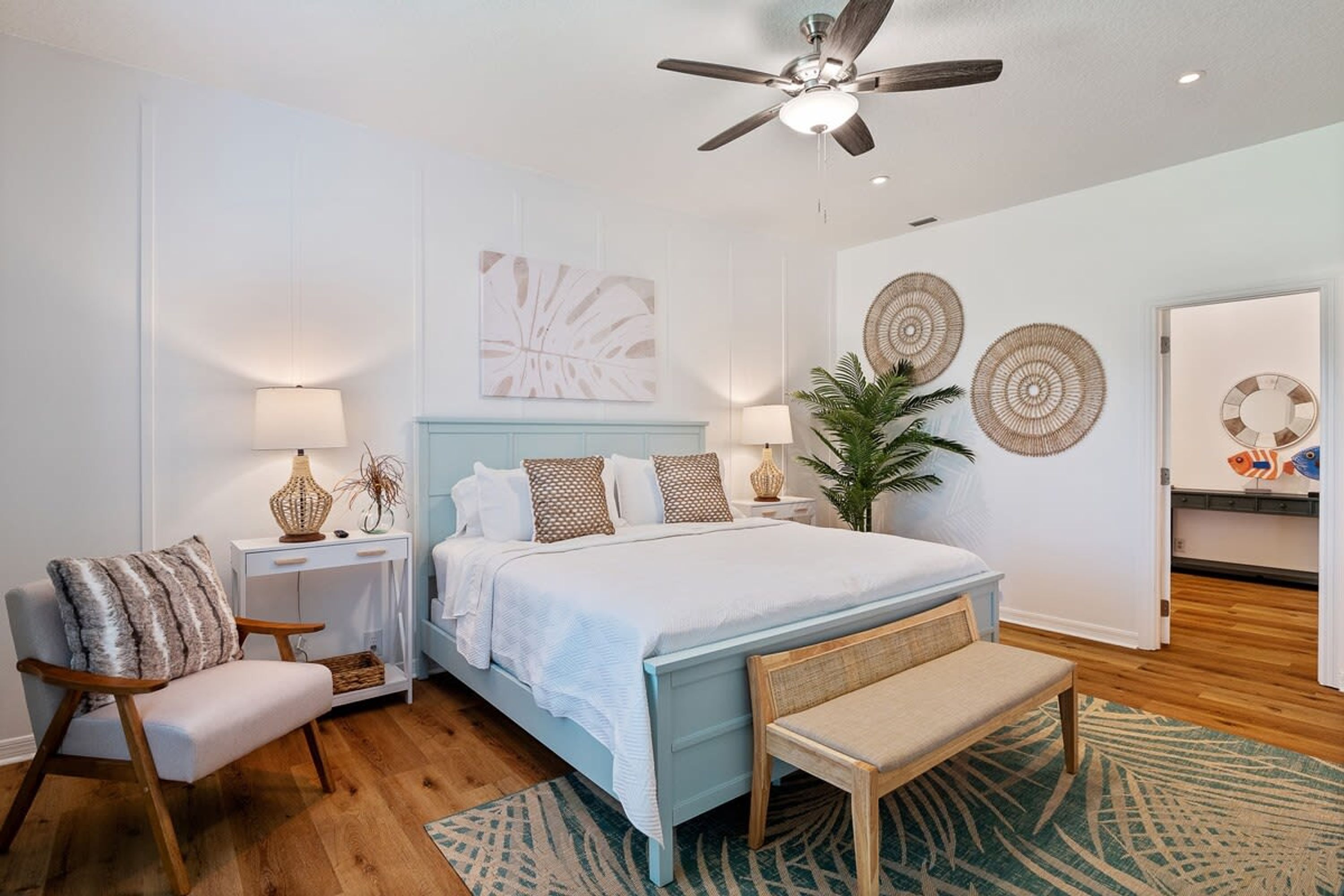 A bedroom with a bed, bedside tables, a chair, a bench, a ceiling fan, wall art, and a door with a fish decoration.