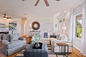 Un salon moderne avec un canapé gris, un canapé blanc et une table basse en bois. On y trouve un ventilateur de plafond, une cheminée et une grande fenêtre avec des rideaux.