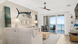 A spacious living room with a large sofa, a ceiling fan, and a view of the ocean through large sliding doors. - New Luxury Ocean Front 3 Bedroom 2 Bath Condo Sleeps 8 Cinnamon Beach Resort (Palm Coast)