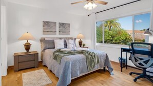Une chambre à coucher comprenant un lit, deux tables de chevet avec des lampes, un bureau avec une chaise, un ventilateur de plafond et une grande fenêtre donnant sur un paysage verdoyant.