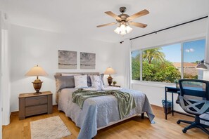 Ein Schlafzimmer mit einem Bett, zwei Nachttischen mit Lampen, einem Schreibtisch mit Stuhl, einem Deckenventilator und einem großen Fenster mit Blick auf Grünflächen.