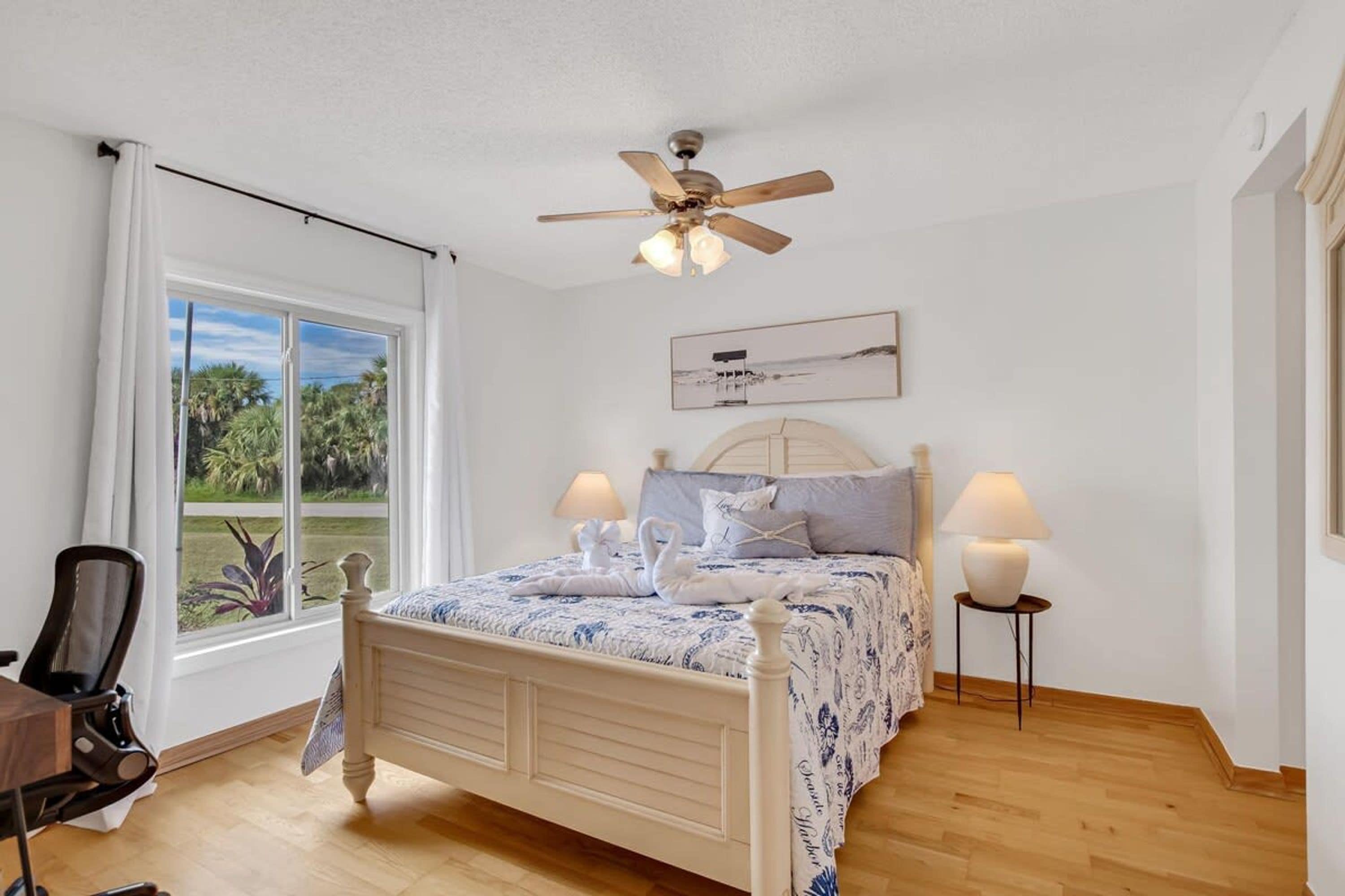 A bedroom with a bed, two bedside lamps, a ceiling fan, a window with curtains, and a chair.