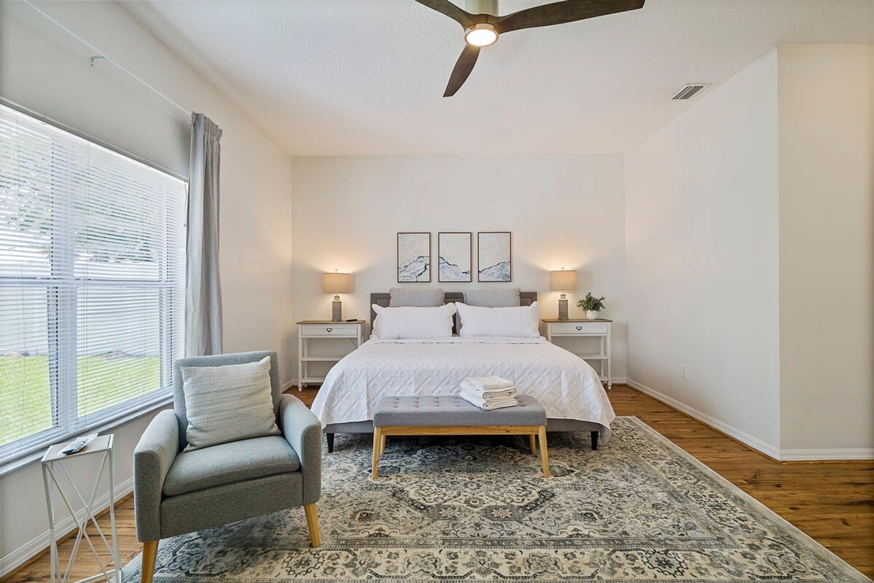 A bedroom with a bed, bedside tables, a chair, a bench, a ceiling fan, and a large window.