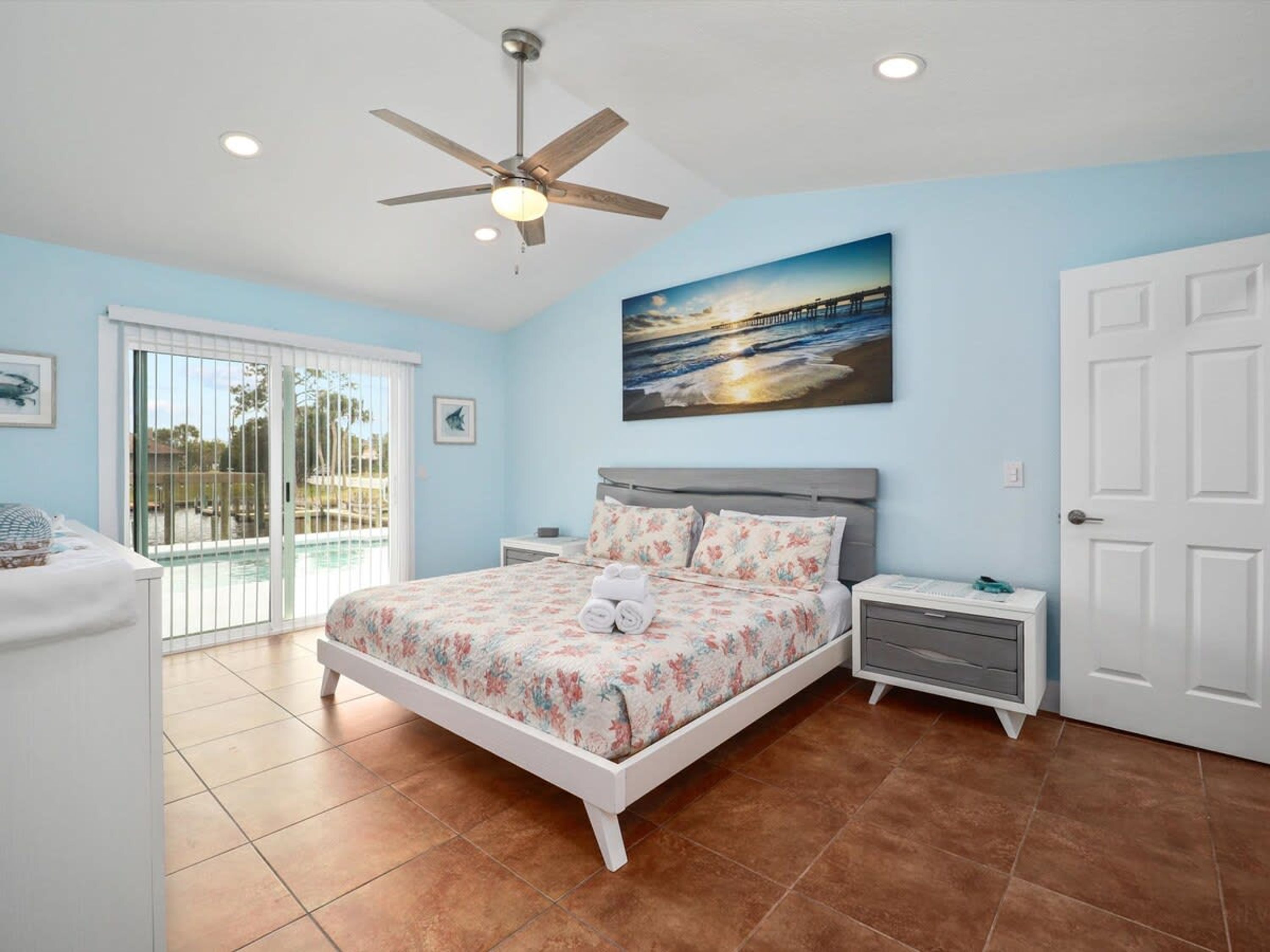 A bedroom with a bed, a nightstand, a ceiling fan, a painting, and a sliding glass door to a pool area.