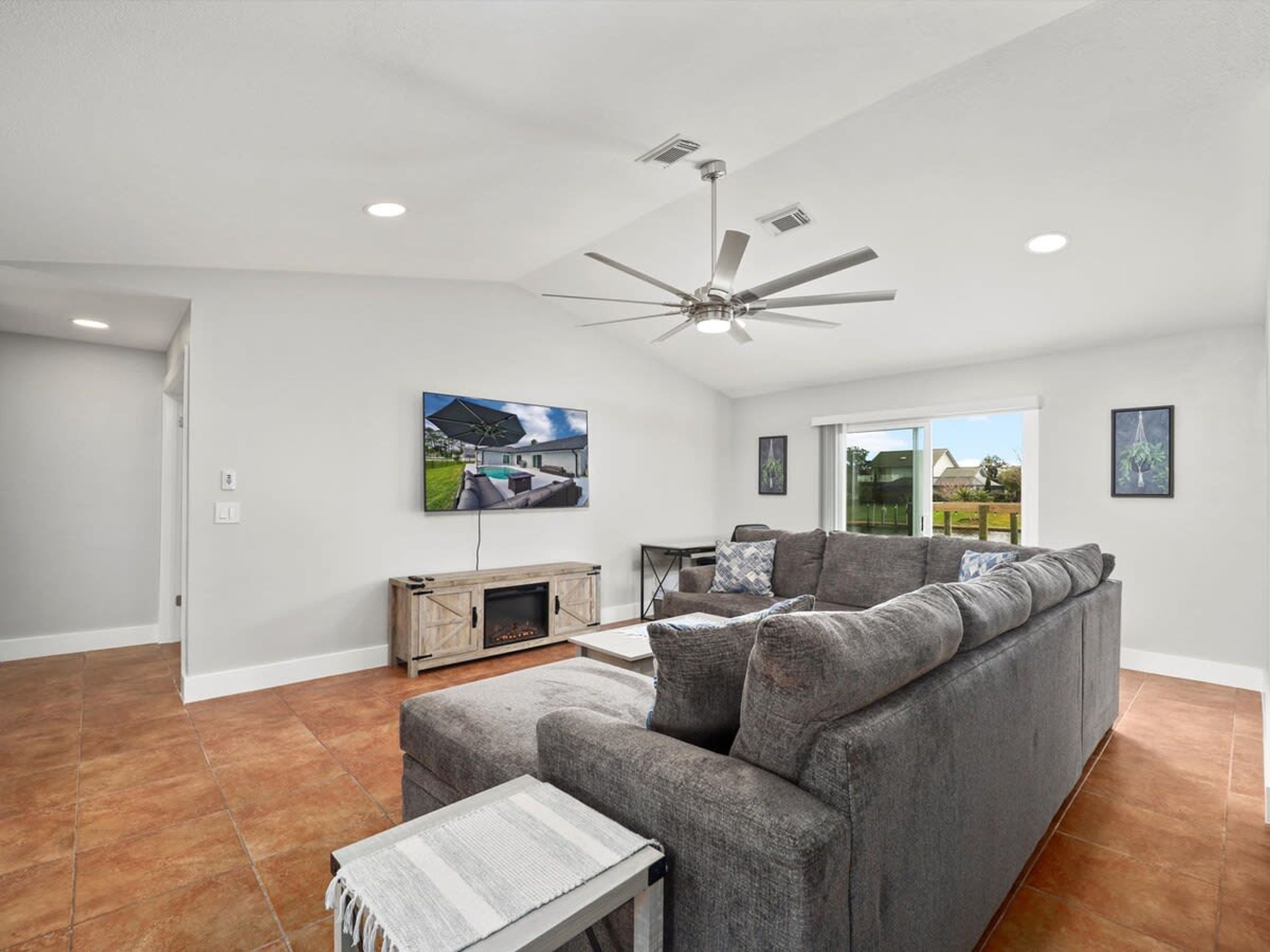 A spacious living room with a grey sectional sofa, a fireplace, a ceiling fan, and a large window offering a view of the outdoors.