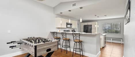 A modern kitchen with a foosball table, bar stools, and a central island with a sink and microwave.