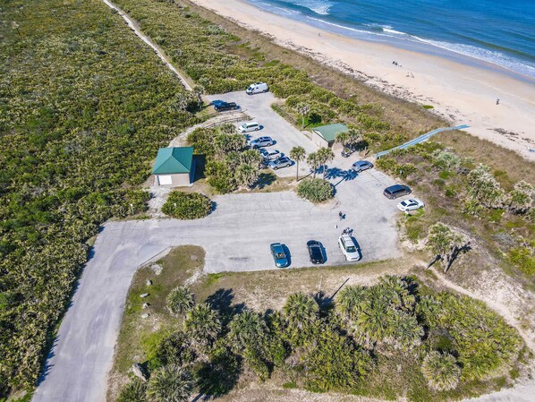 Vue aérienne d’une propriété située sur la côte, avec un parking, un petit bâtiment doté d’un toit végétalisé et une plage en arrière-plan.