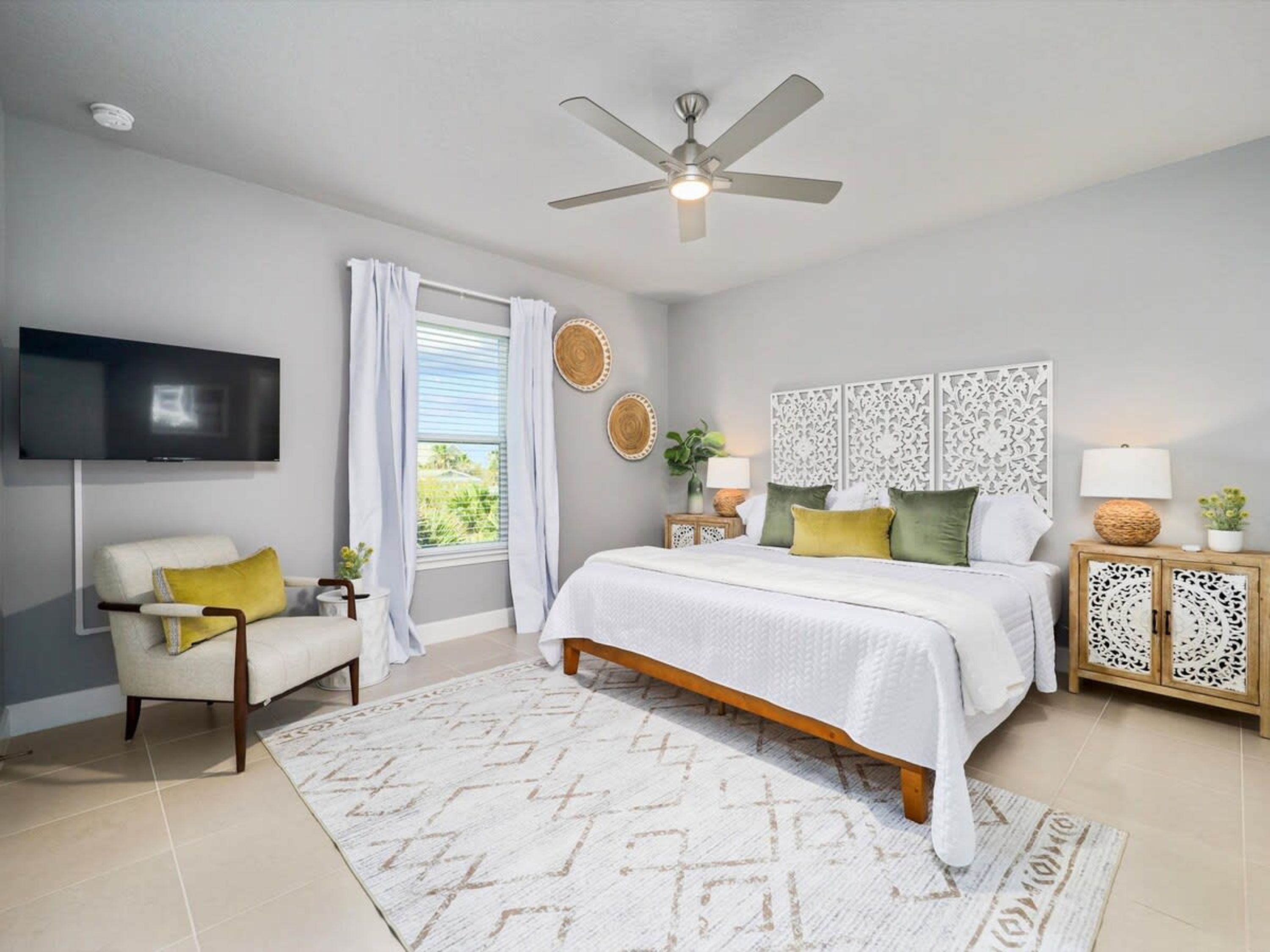Une chambre à coucher comprenant un lit, une chaise, un téléviseur, un ventilateur de plafond et une fenêtre avec des rideaux.