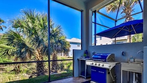 An outdoor kitchen with a grill, sink, and large windows overlooking a garden with palm trees. - Seabliss Steps to the Beach Pool Home 4 4 5 new (Palm Coast)