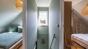 Een compacte, goed verlichte slaapkamer op zolder met een schuine hemel, een klein raam met decoratieve luiken, een bed met een witte dekbedovertrek, een houten trapje en een geweven stoel.