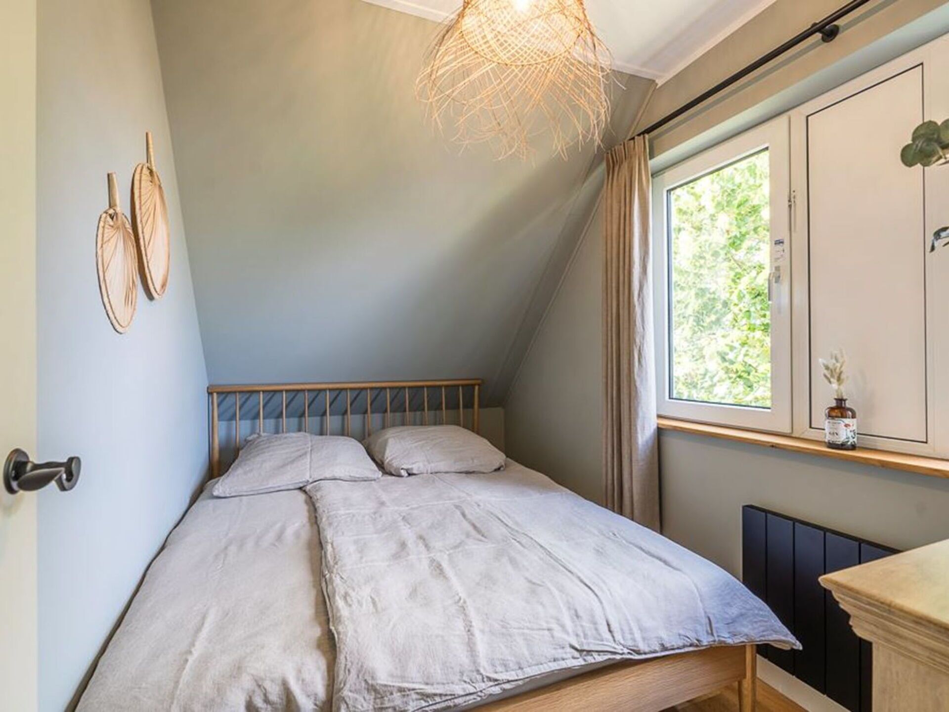 Un dormitorio con techo inclinado, una cama con cabecera de madera, una lámpara colgante y un ventanal con cortinas.