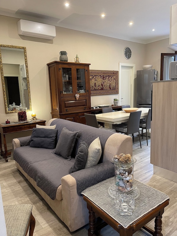 A living room with a sofa, a wooden cabinet, a dining table with chairs, and a kitchen area with a refrigerator.