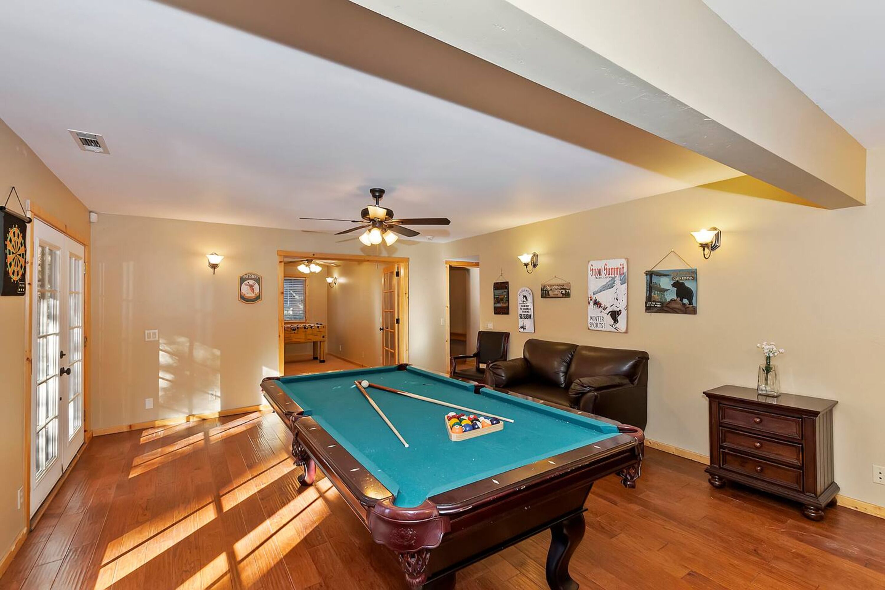 Une table de billard dans une pièce au plancher en bois, avec un ventilateur de plafond et des lampes fixées au mur.