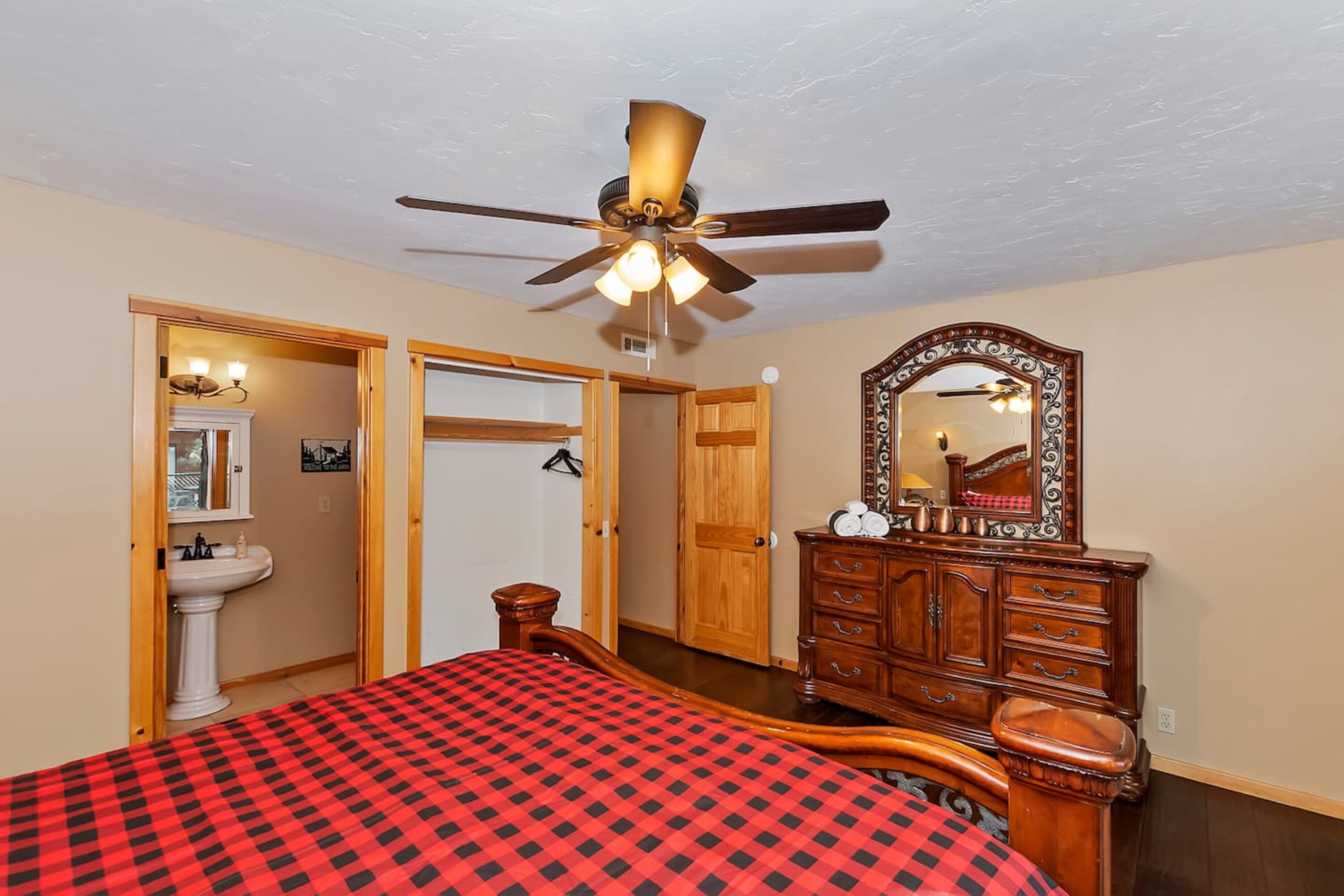 A bedroom with a bed, a wooden dresser, a mirror, a ceiling fan, and a door leading to another room.