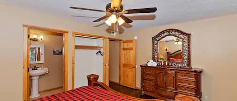 A bedroom with a bed, a wooden dresser, a mirror, a ceiling fan, and a door leading to another room.