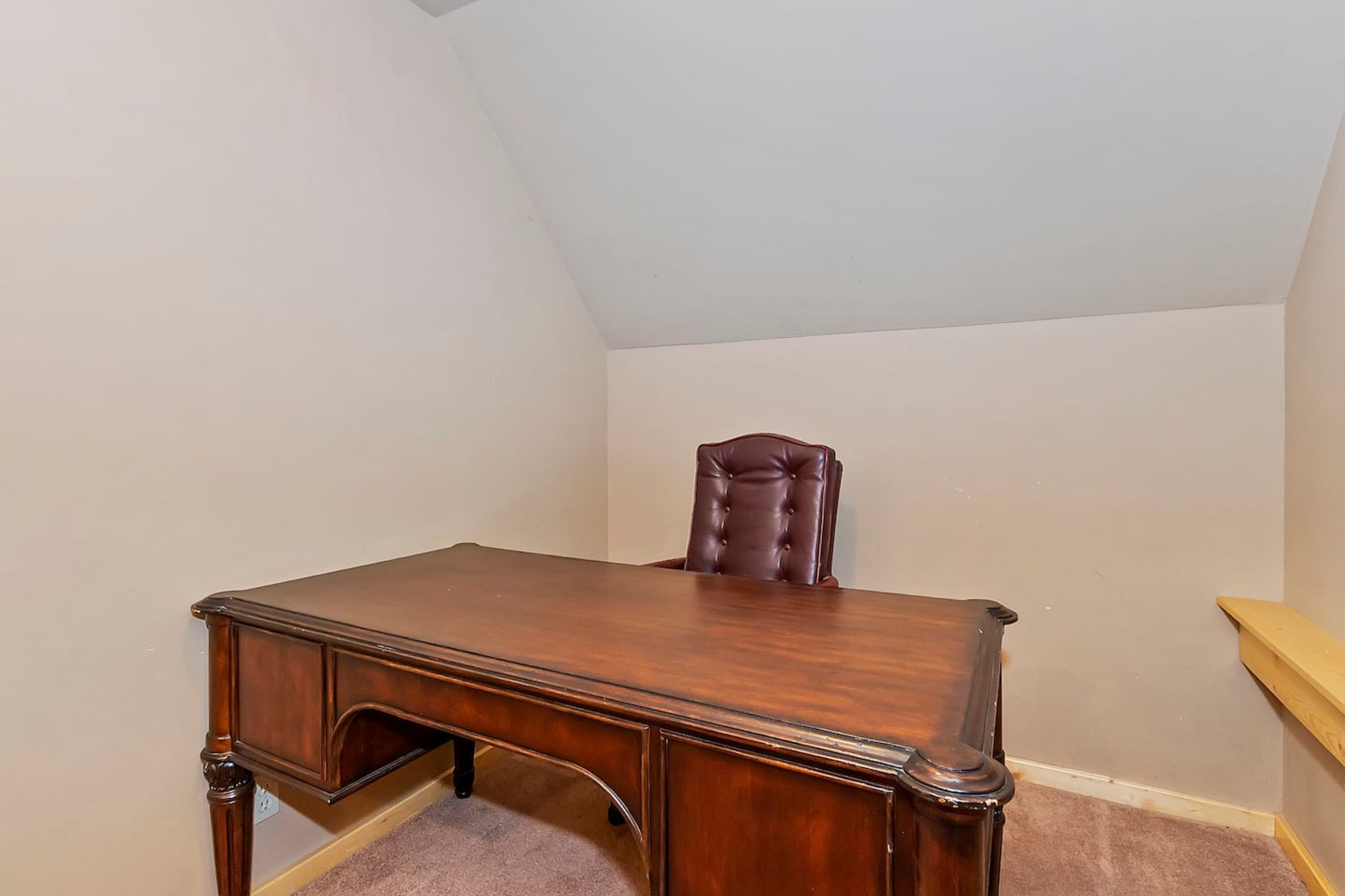 Een houten bureau in een hoek van een kamer met een schuine balkenplafond en een houten stoel.