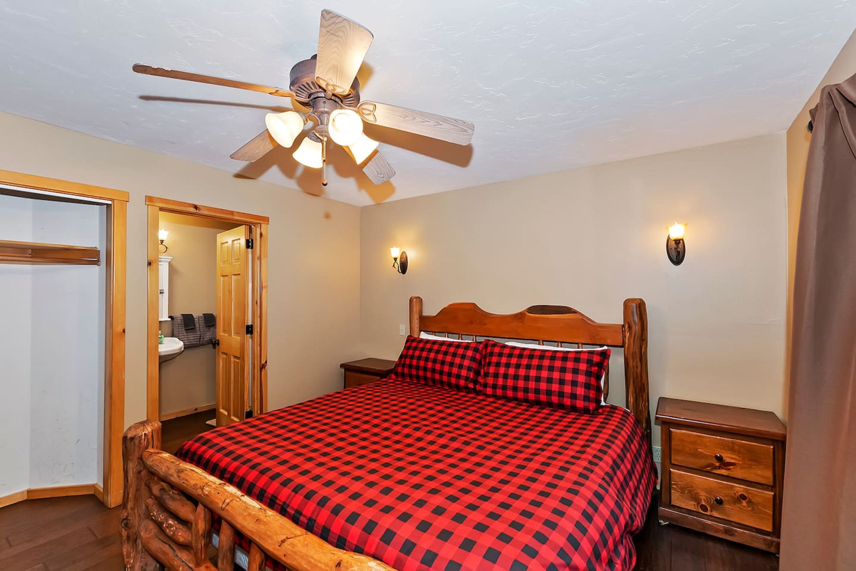 A bedroom with a wooden bed frame, a red and black checkered bedspread, a ceiling fan, two bedside tables, and a wooden wardrobe.