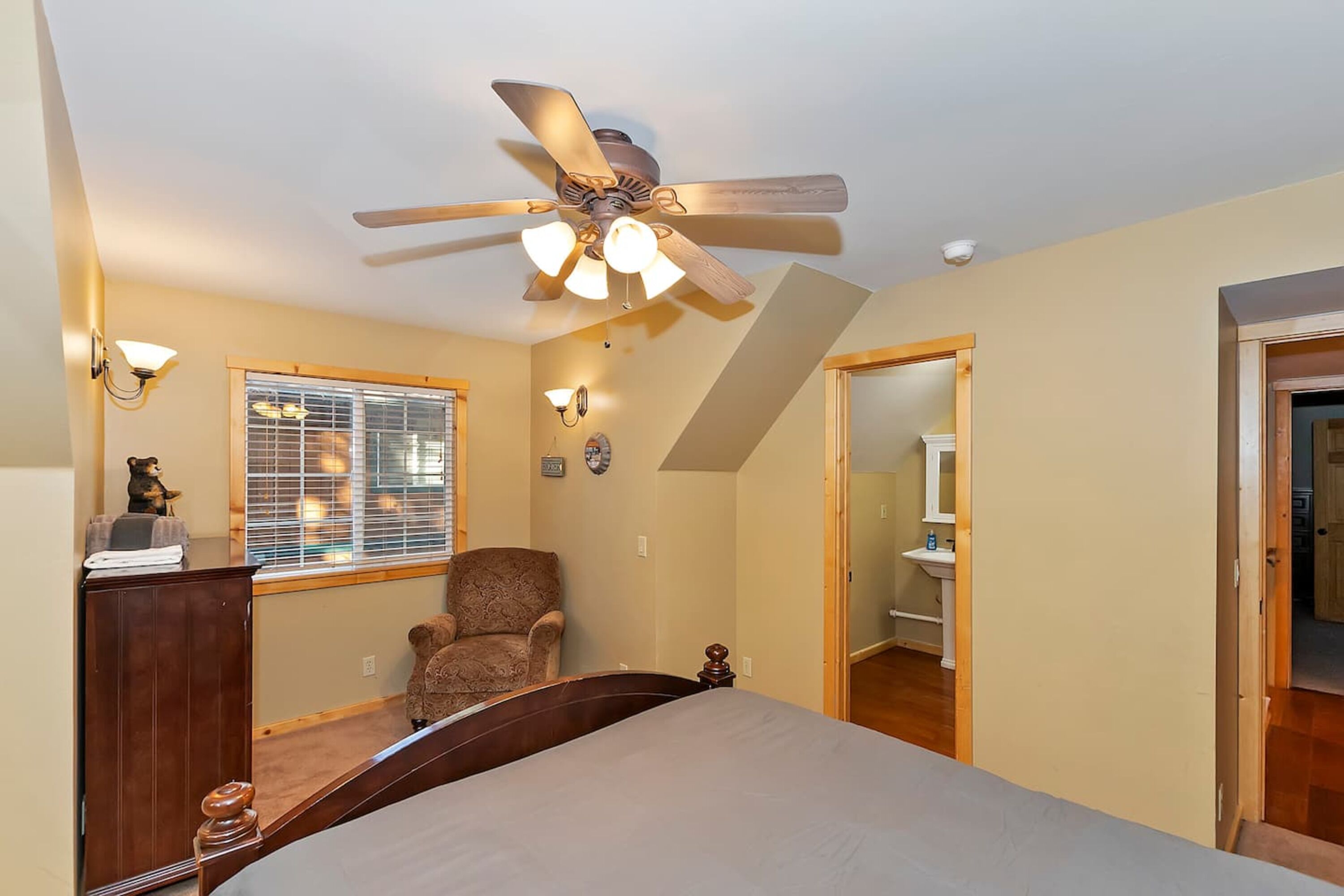 A bedroom with a bed, a chair, a ceiling fan, a window with blinds, and a door leading to a bathroom.