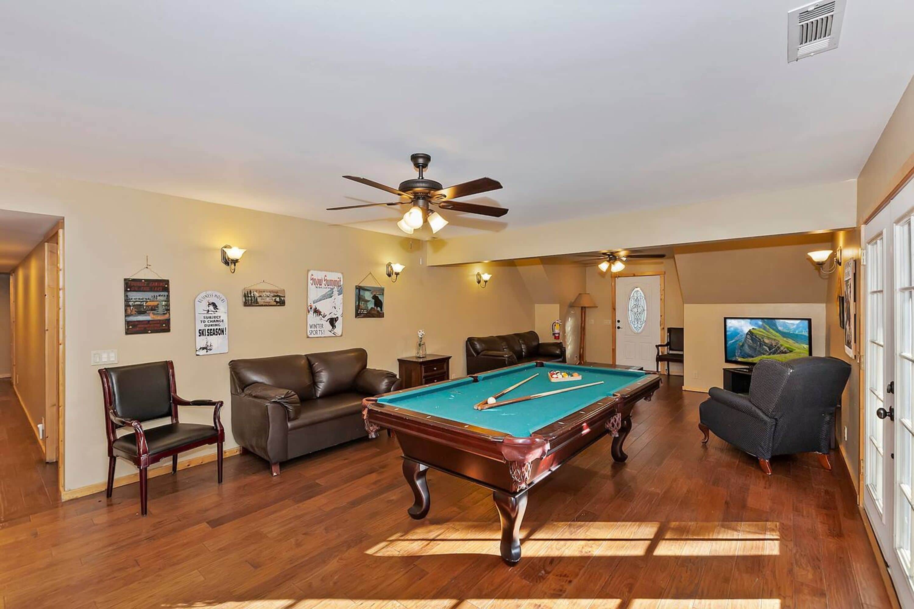 A room with a pool table, two leather sofas, a ceiling fan, and a television.