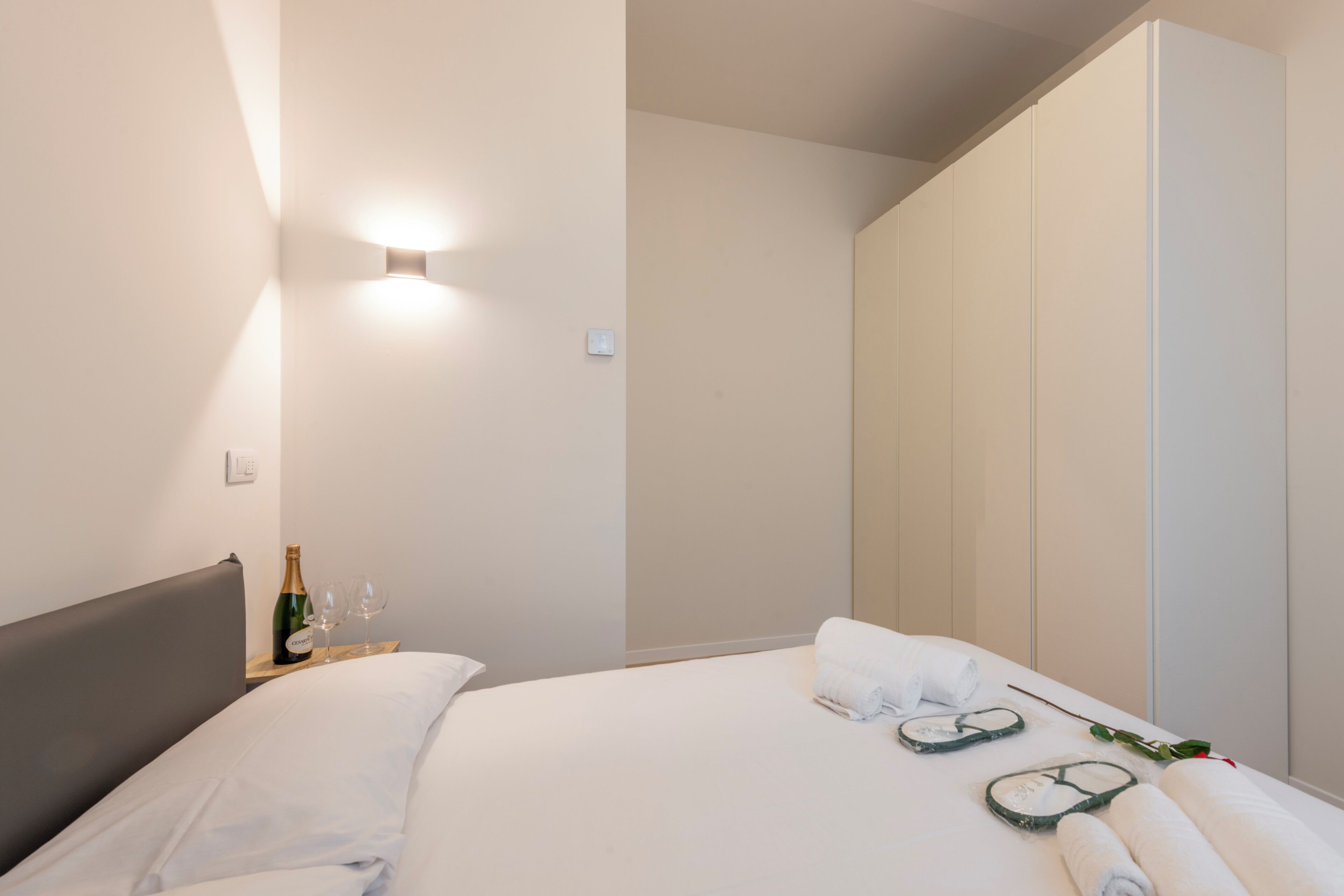 Une chambre d’hôtel moderne avec un lit bien fait, une table de chevet sur laquelle se trouvent une bouteille et des verres, et une grande armoire.