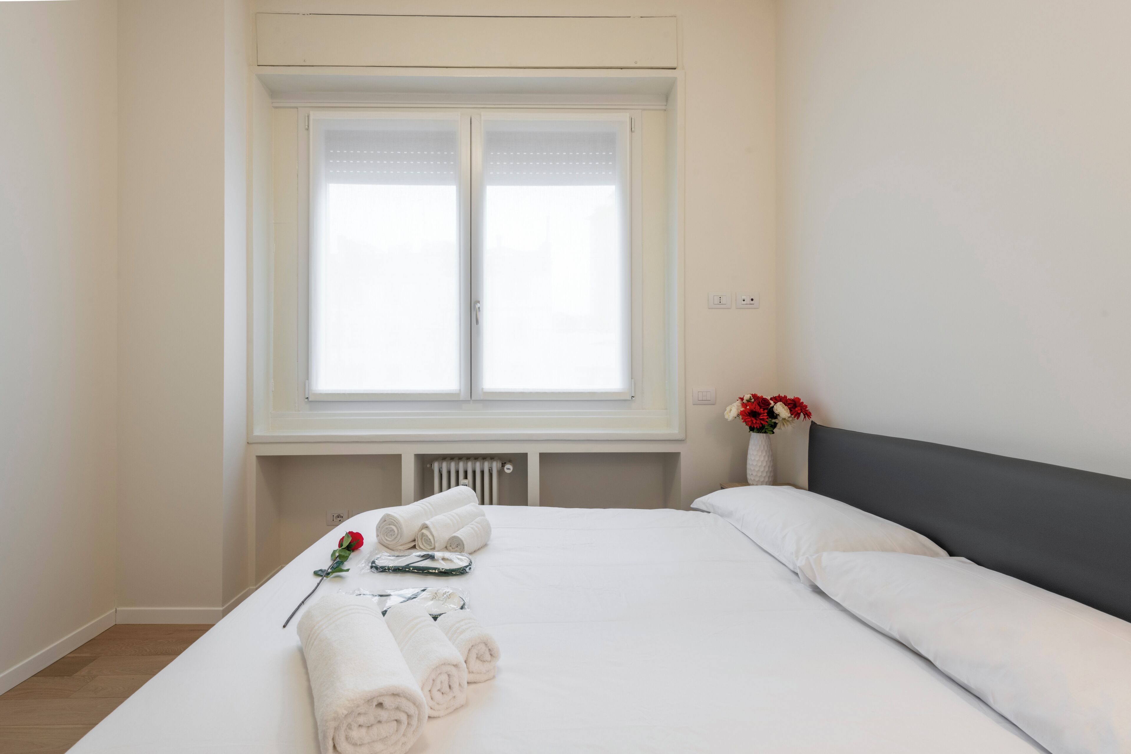 A hotel room with a large bed, a radiator below the window, and a vase with flowers on the nightstand.