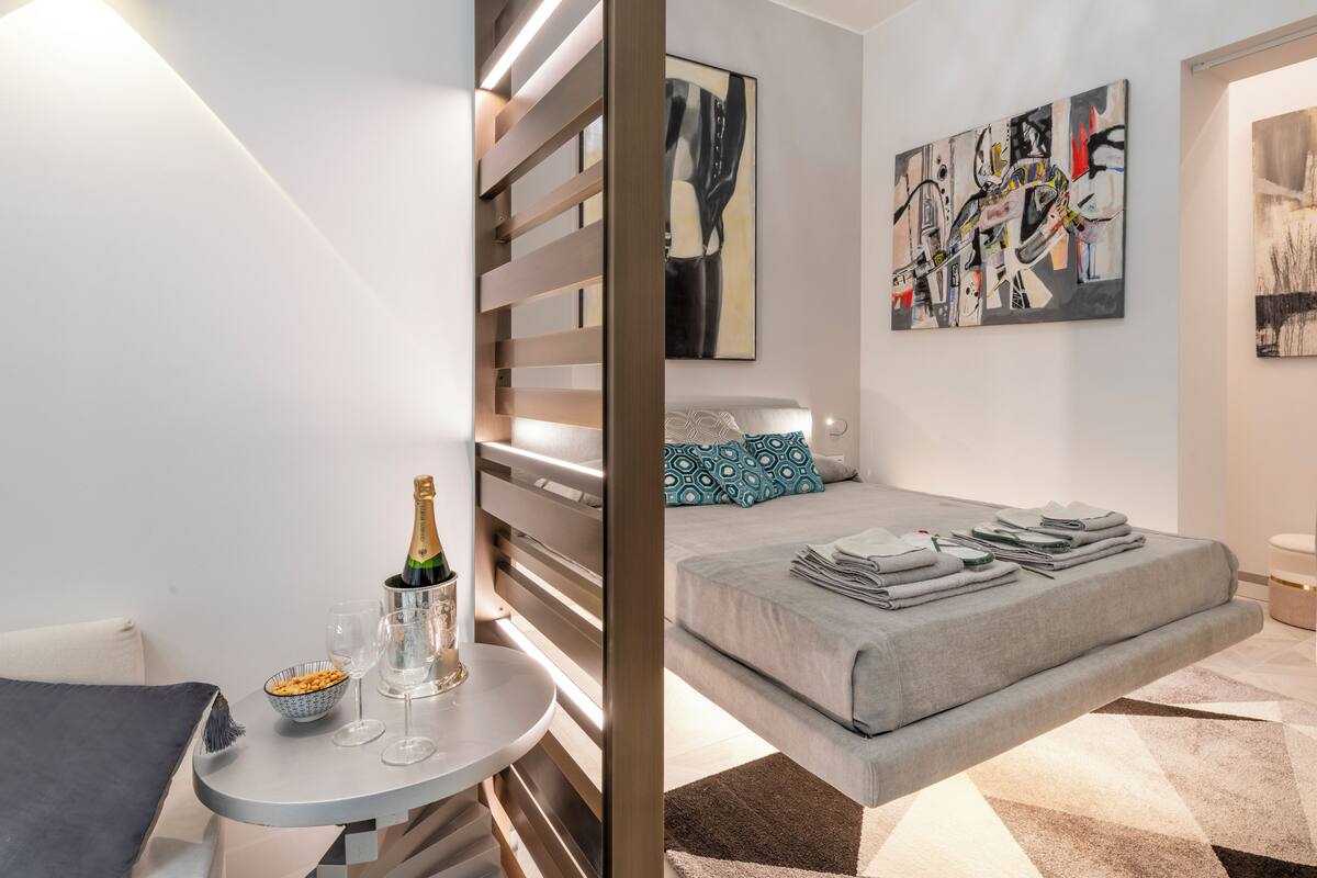 A modern hotel room with a bed, a small table with a bottle of champagne and glasses, and abstract art on the walls.