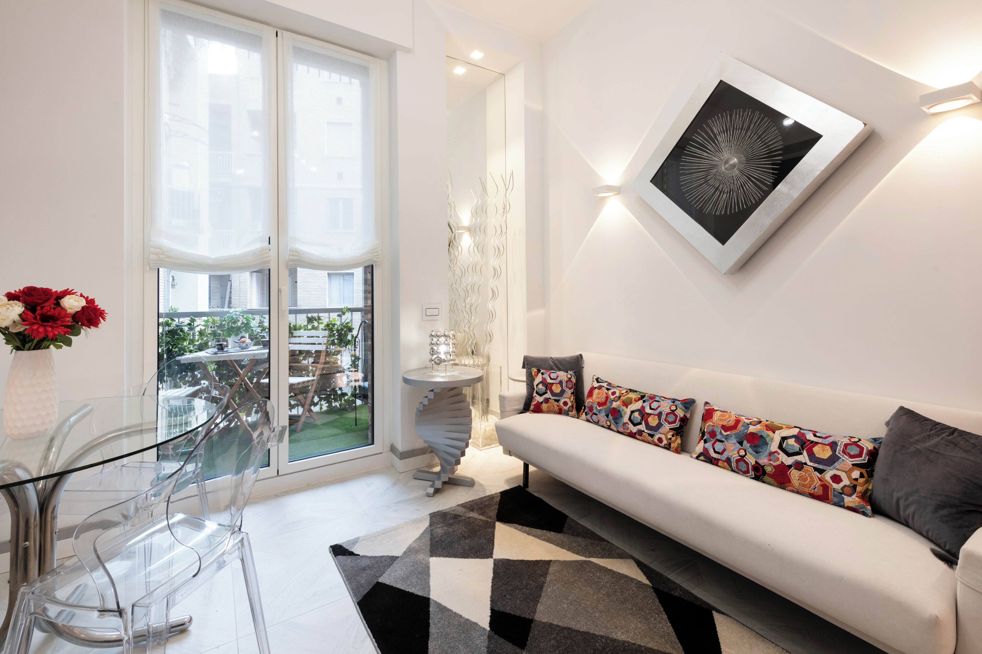 A modern living room with a glass dining table, a white sofa with colorful cushions, a large window, and a decorative wall piece.
