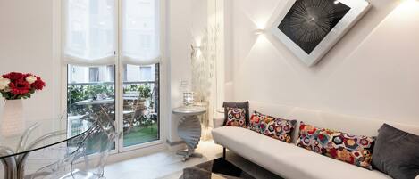 A modern living room with a glass dining table, a white sofa with colorful cushions, a large window, and a decorative wall piece.