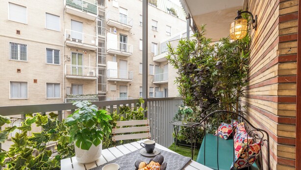 Un balcón con una mesa lista para dos personas, una planta en maceta y una zona de estar con cojines estampados.