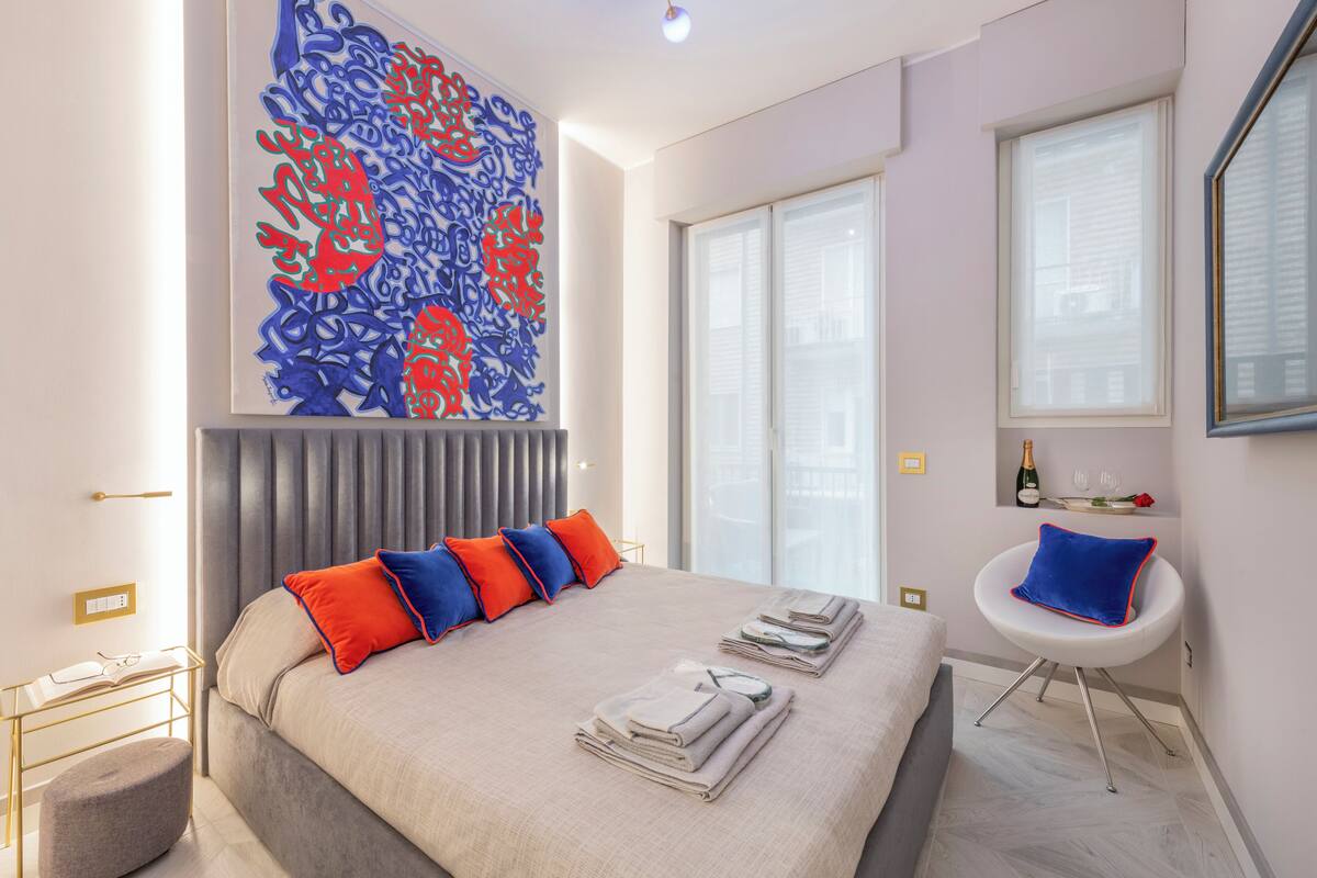 A modern hotel room with a large bed, a seating area with a white chair, a large abstract painting, and a window with blinds.