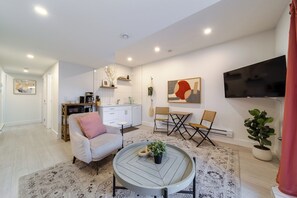 Un salon moderne avec un canapé, une table basse et des fauteuils, un coin cuisine doté de meubles blancs et un téléviseur à écran plat fixé au mur.