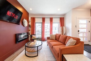 Un salon comprenant un canapé, une table basse, un téléviseur fixé au mur et une fenêtre avec des rideaux.