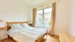 A bedroom with a wooden bed, a nightstand with a lamp, a window with curtains, and a view of buildings outside. - Unique by Grand Apartments (Sopot)