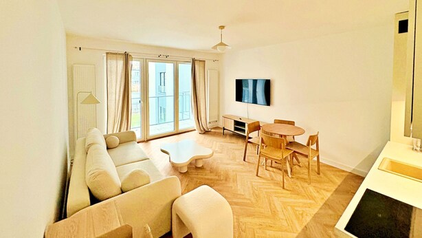 A modern living room with a sofa set, a coffee table, a dining table with chairs, a flat-screen TV mounted on the wall, and large sliding glass doors leading to an outdoor balcony. - Unique by Grand Apartments (Sopot)
