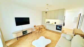 A modern living room with a flat-screen TV, a round dining table with chairs, a white sofa, and a kitchen area with cabinets and a stove. - Unique by Grand Apartments (Sopot)