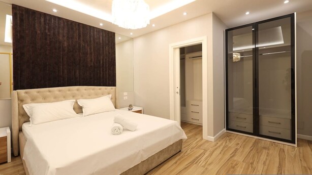 Een moderne slaapkamer met een groot bed, een wandpaneel van donker hout, een inloopkast en een kroonluchter.