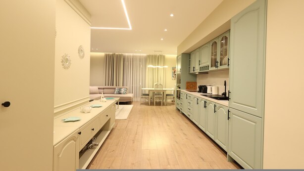Una cocina moderna con armarios verde claro, una zona de comedor con una mesa y sillas, y un sofá al fondo.
