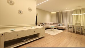 Una sala de estar moderna con un sofá color beige, una mesa de comedor con sillas, un aparador blanco con cajones y apliques decorativos.