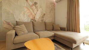 En moderne stue med en beige modulsofa, et træsofabord og et stort vægmaleri med tropiske blade.