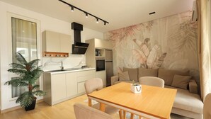 Et moderne køkken med hvide skabe, en indbygget ovn og et sort emfang. Der er et spiseområde med et træbord og stole, samt en beige sofa med en væg bagved i et tropisk blad-mønster.
