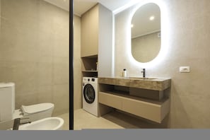A modern bathroom with a built-in washing machine, a sink with a marble countertop, and a large mirror with integrated lighting. - Grandstay Apartments 001 (Durrës)