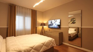 A hotel room with a large bed, a TV mounted on the wall, a lamp, a dresser, and a mirror reflecting the bed and a painting on the wall. - Grandstay Apartments 001 (Durrës)