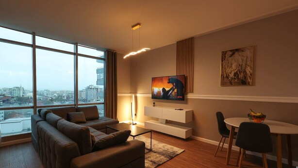 A modern living room with a large window, a sofa, a TV, a dining table, and a painting. - Grandstay Apartments 001 (Durrës)
