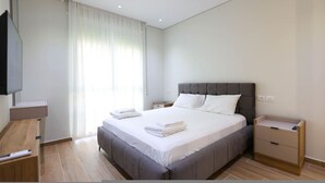 Ein modernes Schlafzimmer mit einem großen Bett, einem Nachttisch, einem an der Wand befestigten Fernseher und einem Fenster mit durchsichtigen Vorhängen.