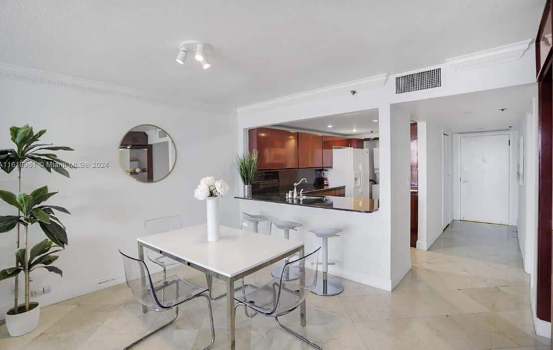 A modern kitchen with white cabinets, a black countertop, and a white refrigerator. There is a dining area with a white table and chairs, and a potted plant.