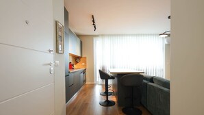 Een compacte keuken met een eethoek, met een houten vloer, een grijze bank en een kleine tafel met twee krukken.