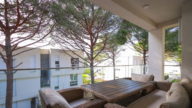 Een balkon met rieten meubels, een houten tafel en uitzicht op gebouwen en bomen.