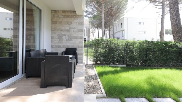 A modern outdoor patio with wicker furniture, a stone wall, and a well-maintained grassy area with a hedge and trees in the background. - Sanpietro Vacation Apartment 009 (Ishëm)