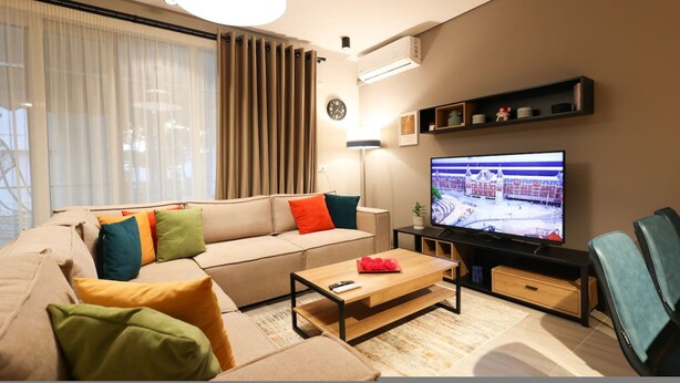 A modern living room with a beige sofa, colorful cushions, a wooden coffee table, a flat-screen TV, and a large window with sheer curtains. - Sanpietro Vacation Apartment 011 (Ishëm)